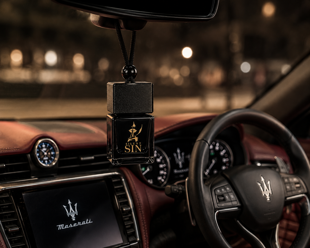 A black car air freshener or fragrance bottle labeled ' SILENT SIN' with a gold trident logo, hanging from the rearview mirror inside a luxury car cabin with a visible Maserati steering wheel and dashboard. Silent Sin Automotive Elixir Car Fragrance