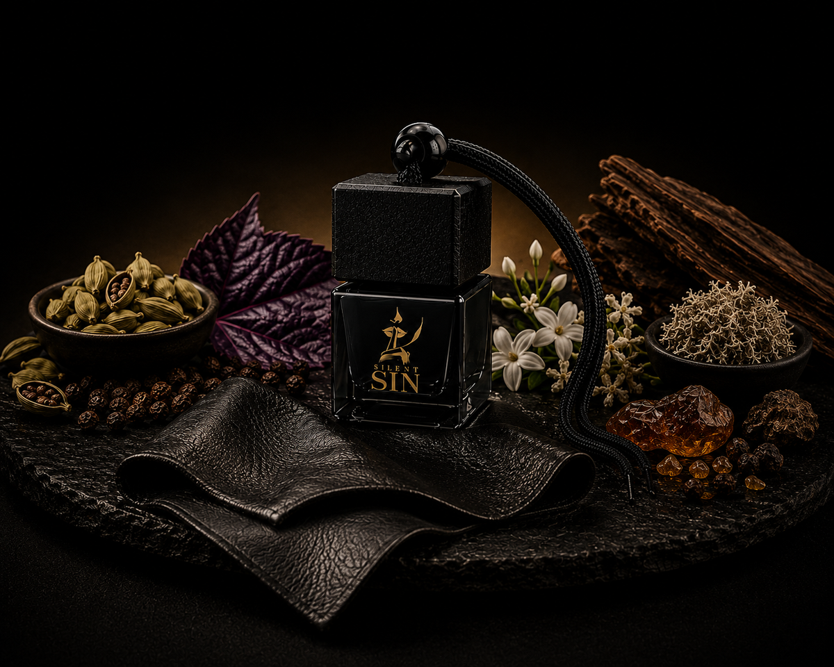 A black rectangular perfume bottle labeled 'SILENT SIN' with a black leather neck strap, styled on a dark stone surface with leather, spices, flowers, and dried botanicals against a black background. Silent Sin Automotive Elixir Car Fragrance