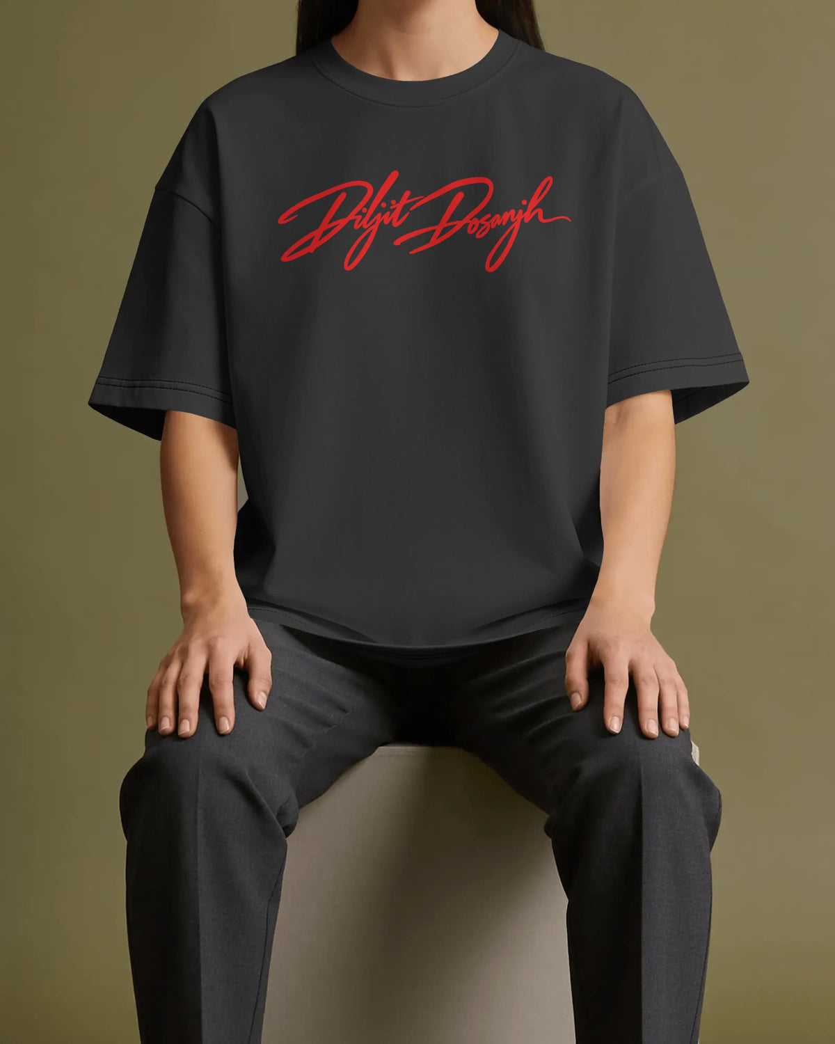 A person seated against a muted olive background wearing an oversized dark charcoal T-shirt with a red script logo across the chest and dark pants. DIljit Dosanjh ura Tour Tshirt