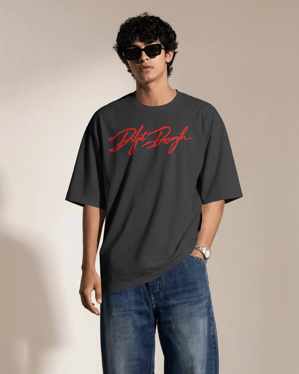A man wearing a dark charcoal gray t-shirt with a red 'Diljit Dosanjh' script logo on the chest, paired with blue jeans and black sunglasses, standing against a neutral beige background.