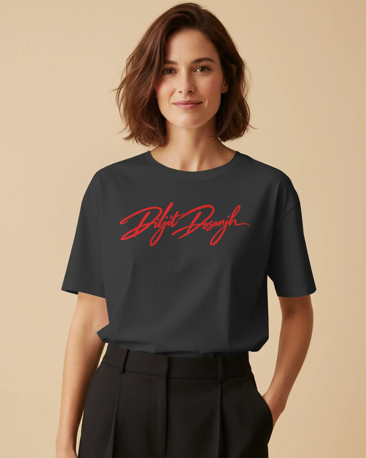 A woman wearing a dark charcoal gray t-shirt with the words 'Diljit Dosanjh' printed in red cursive across the chest, paired with black trousers, set against a solid beige background.