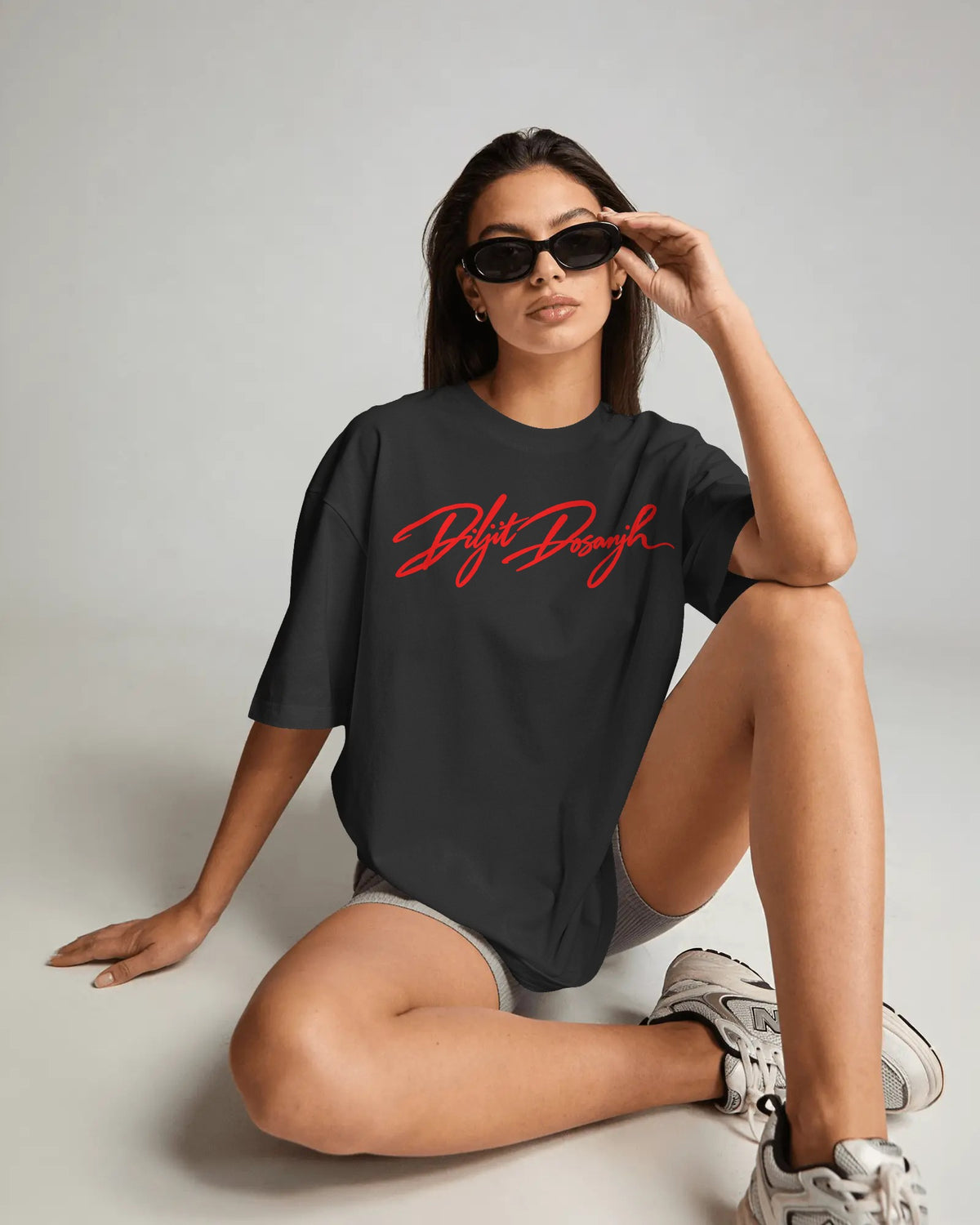 A woman wearing a black oversized t-shirt with a red 'Diljit Dosanjh' script logo across the chest, paired with gray shorts and white sneakers, posing on a light gray studio floor.