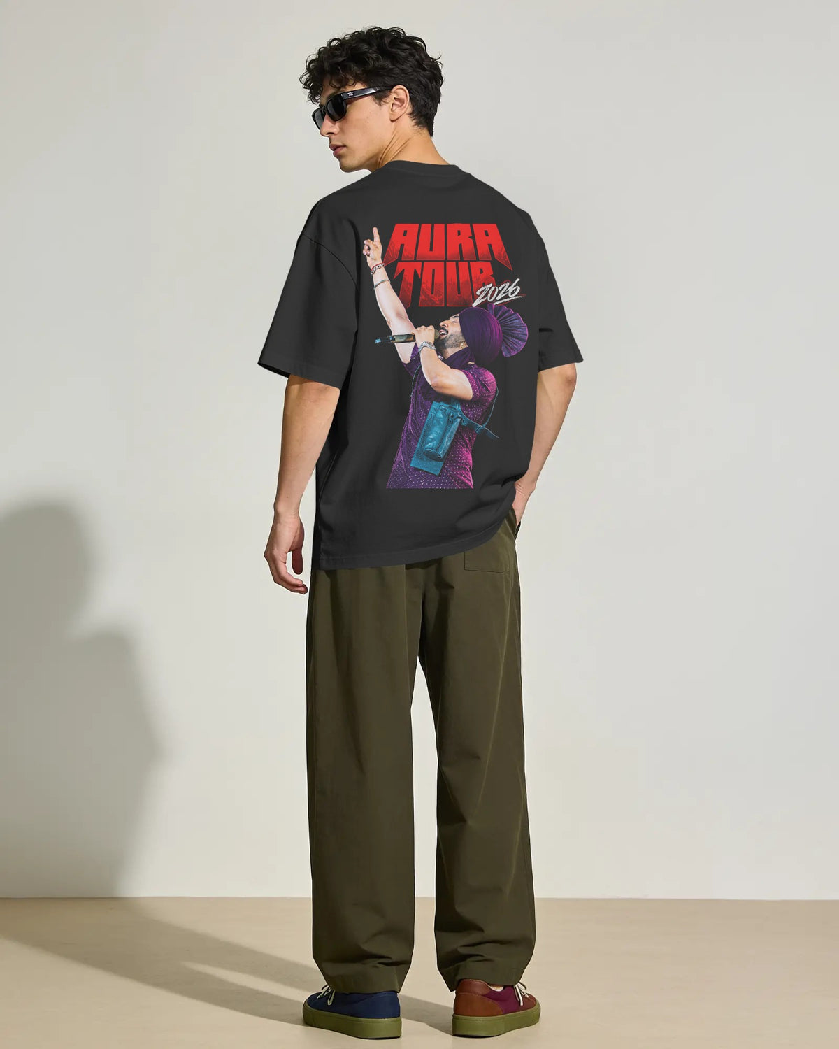 A man wearing a black graphic t-shirt with a large ' Diljit Dosanjh AURA TOUR 2026 merchandise' print on the back, paired with olive green pants and sunglasses, standing in a studio setting. 