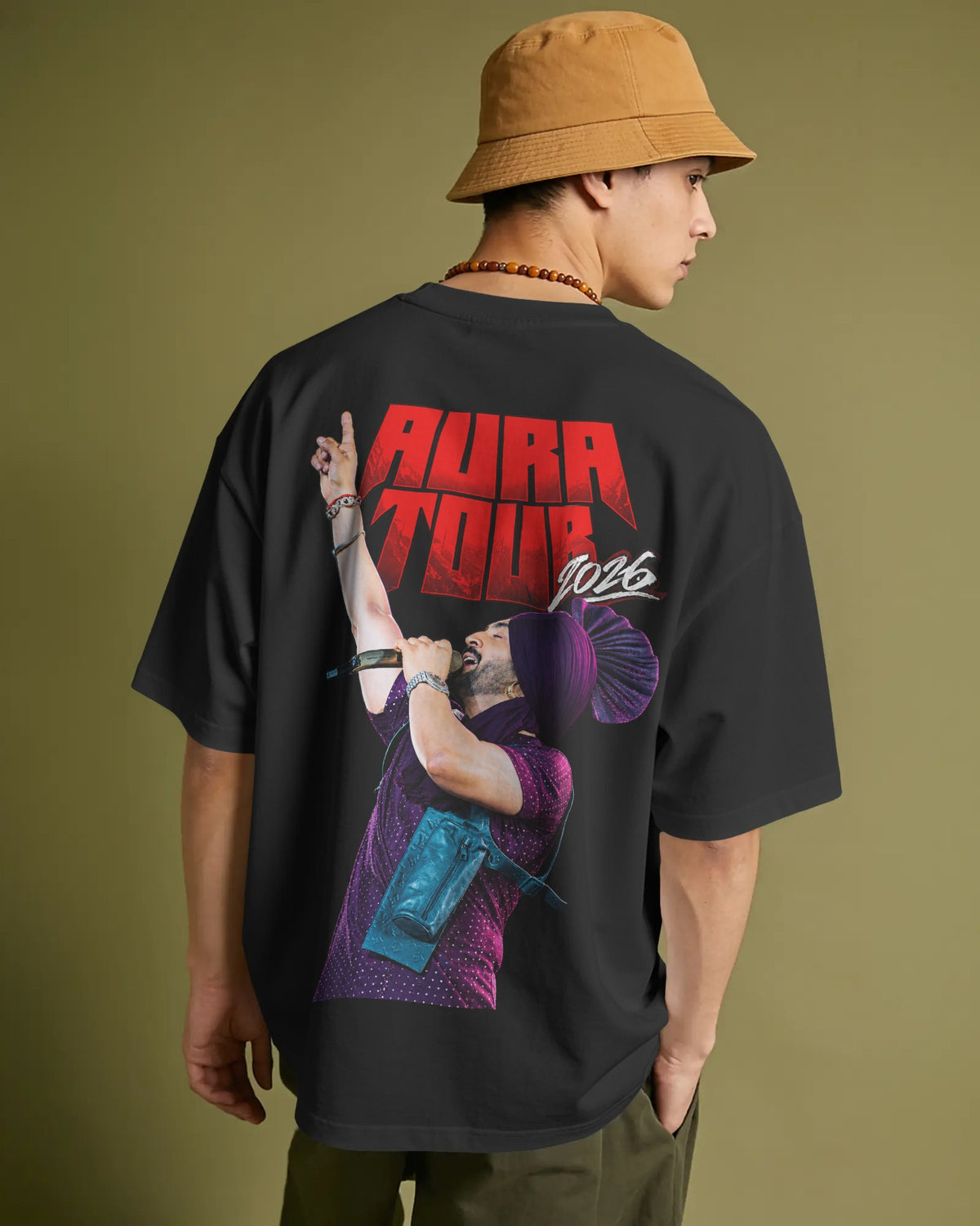 A man wearing a black oversized t-shirt with a large graphic print on the back, featuring red 'AURA TOUR 2026' text and a purple-toned concert illustration. He is also wearing a tan bucket hat and olive pants, photographed against a solid olive green studio background. Diljit Dosanjh Aura Tour India Tshirt