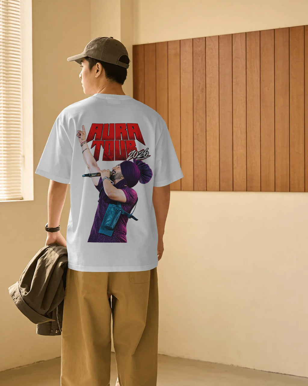 A man wearing a white Diljit Dosanjh Aura Tour India t-shirt with a large red and purple graphic print on the back, paired with tan pants and a brown cap, standing in a room with a wood-paneled wall. 