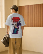 A man wearing a white Diljit Dosanjh Aura Tour India t-shirt with a large red and purple graphic print on the back, paired with tan pants and a brown cap, standing in a room with a wood-paneled wall. 