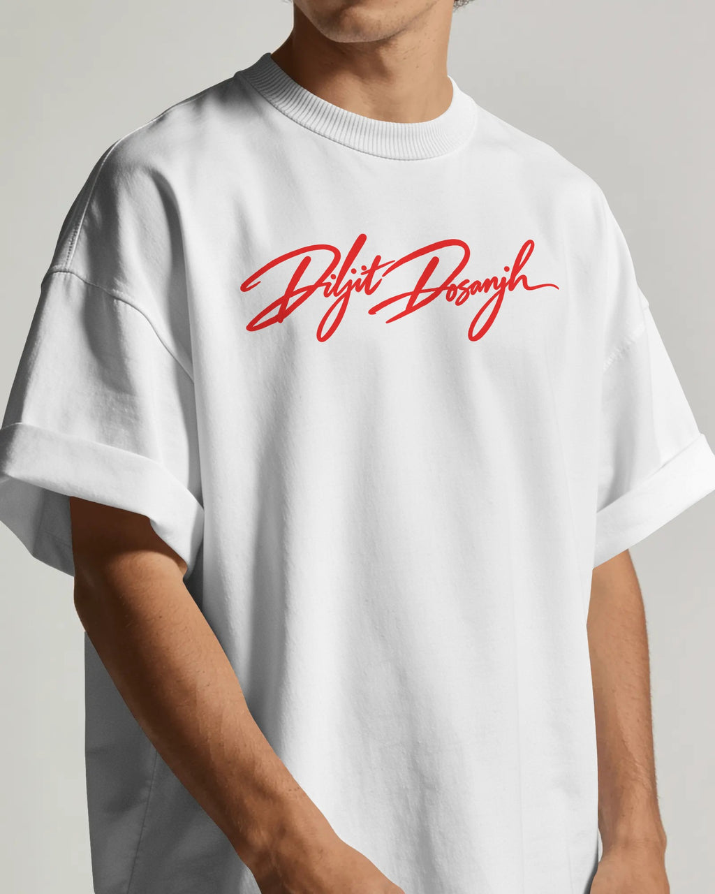A person wearing an oversized white crewneck T-shirt with a red cursive 'Diljit Dosanjh Aura Tour India Tshirt' graphic printed across the chest, shown against a neutral light gray background.