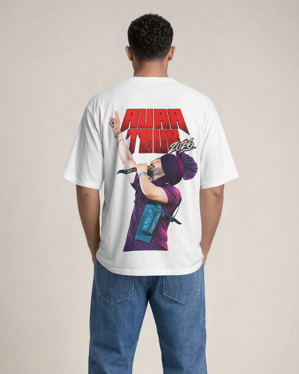 A man viewed from behind wearing a white t-shirt with a large graphic print on the back featuring the text 'AURA TOUR' in red and a purple-haired performer, paired with blue jeans, against a plain beige background.