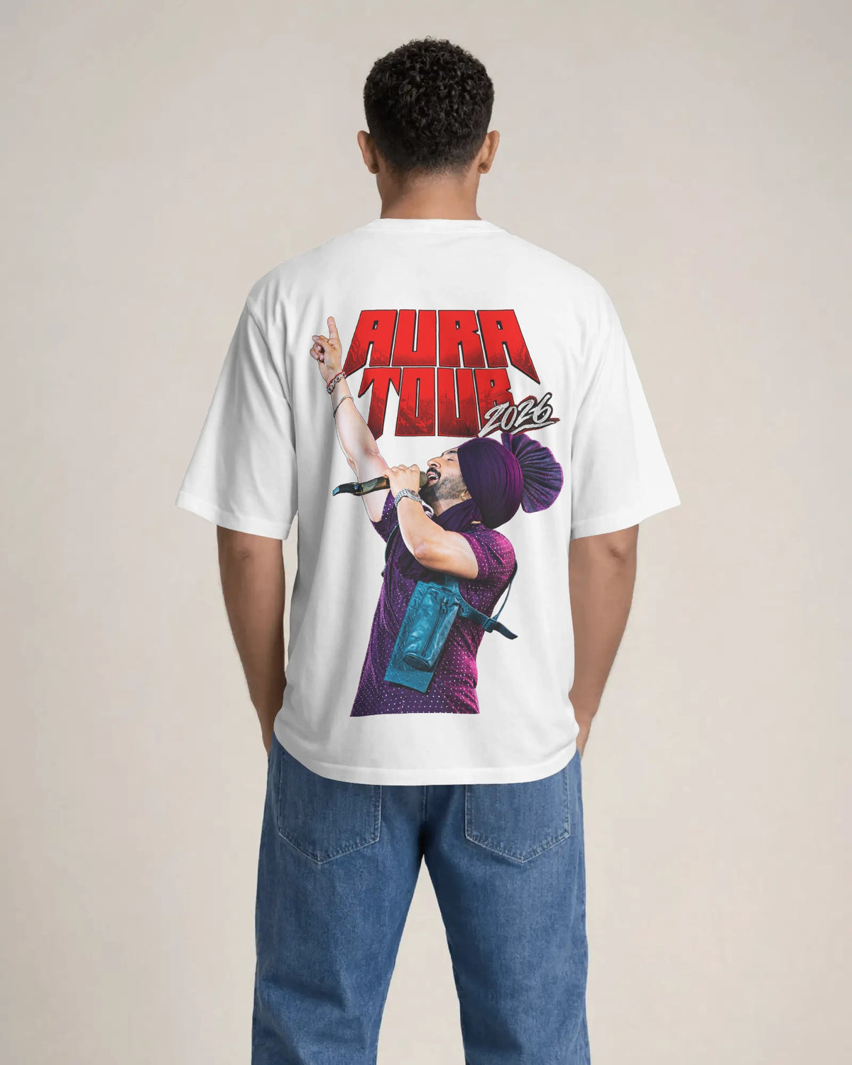 A man viewed from behind wearing a white t-shirt with a large graphic print on the back featuring the text 'AURA TOUR' in red and a purple-haired performer, paired with blue jeans, against a plain beige background.