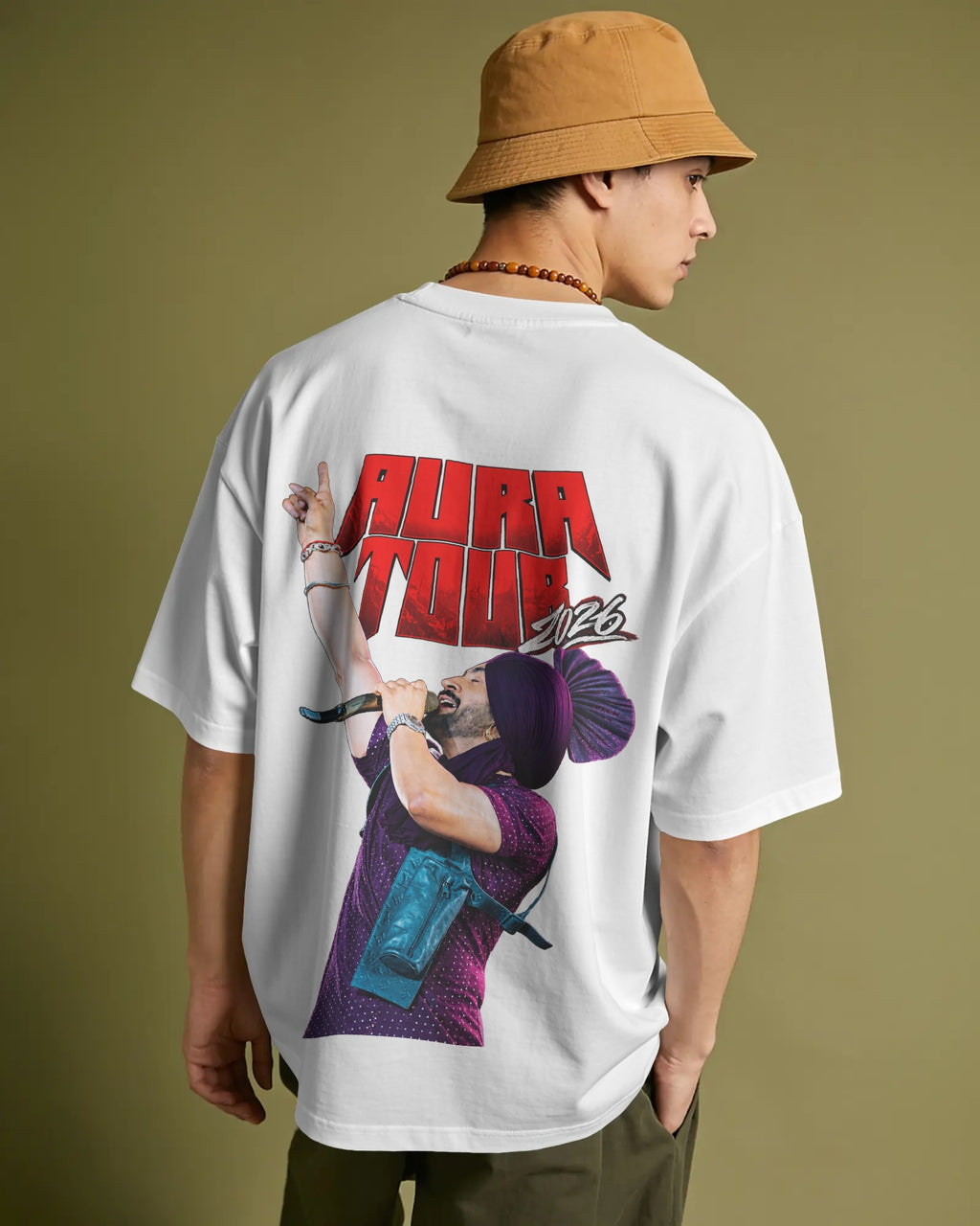 A man wearing a white oversized t-shirt with a large graphic print on the back, featuring the text 'AURA TOUR 2026' and an illustration of a person holding a microphone. He is also wearing a tan bucket hat and olive green pants, photographed against a solid olive green background.