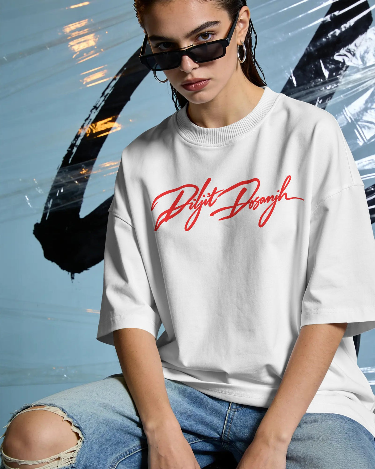 A woman wearing an oversized white crewneck t-shirt with a red cursive graphic print on the chest, paired with ripped blue jeans and black sunglasses, posed against a blue background with a black brushstroke graphic. Diljit Dosanjh Aura Tour India Tshirt