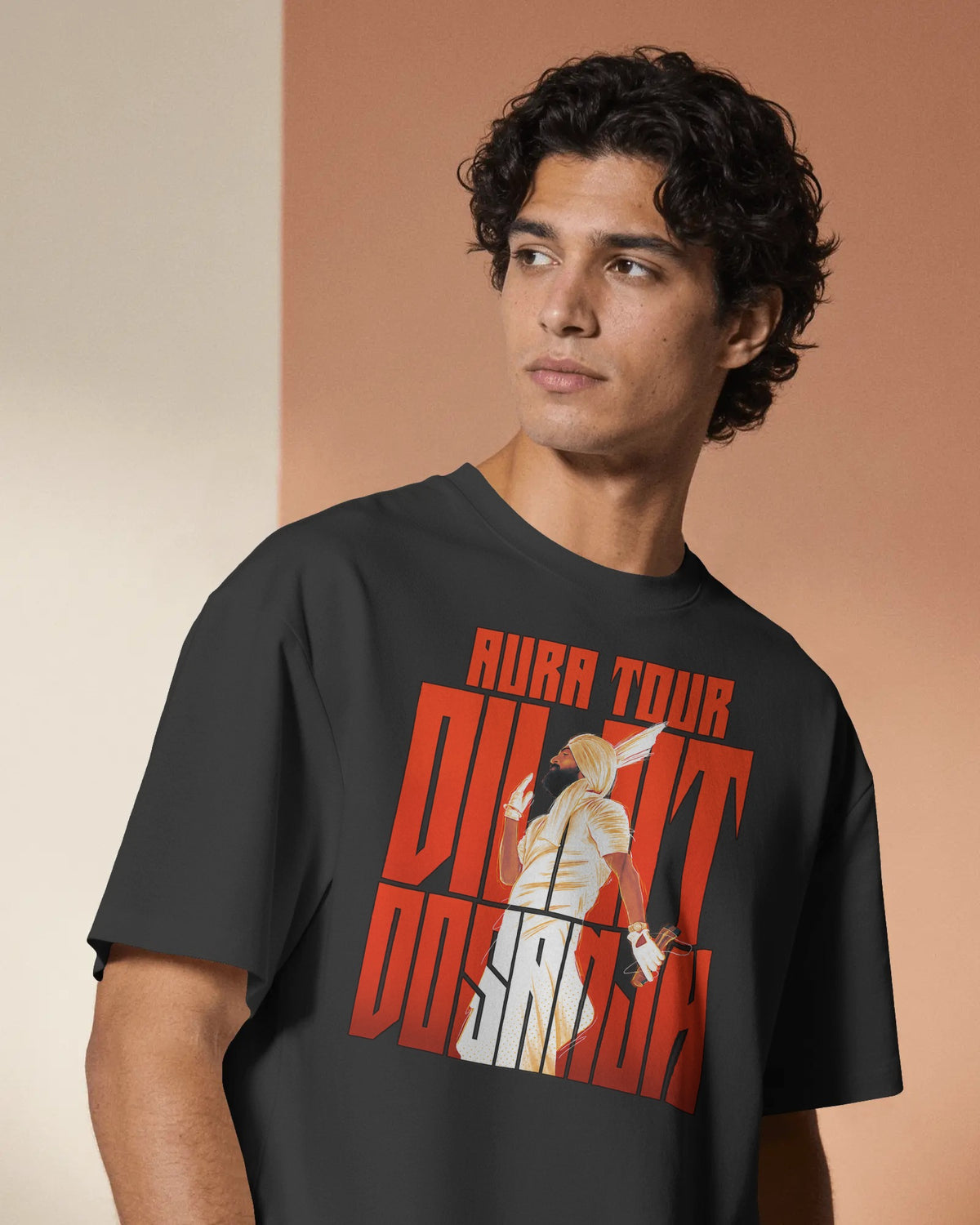 A man wearing a dark charcoal gray t-shirt with a bold red, white, and beige graphic print on the front that reads 'AURA TOUR Diljit Dosanjh'. The background is a split-color studio wall in cream and warm terracotta tones.