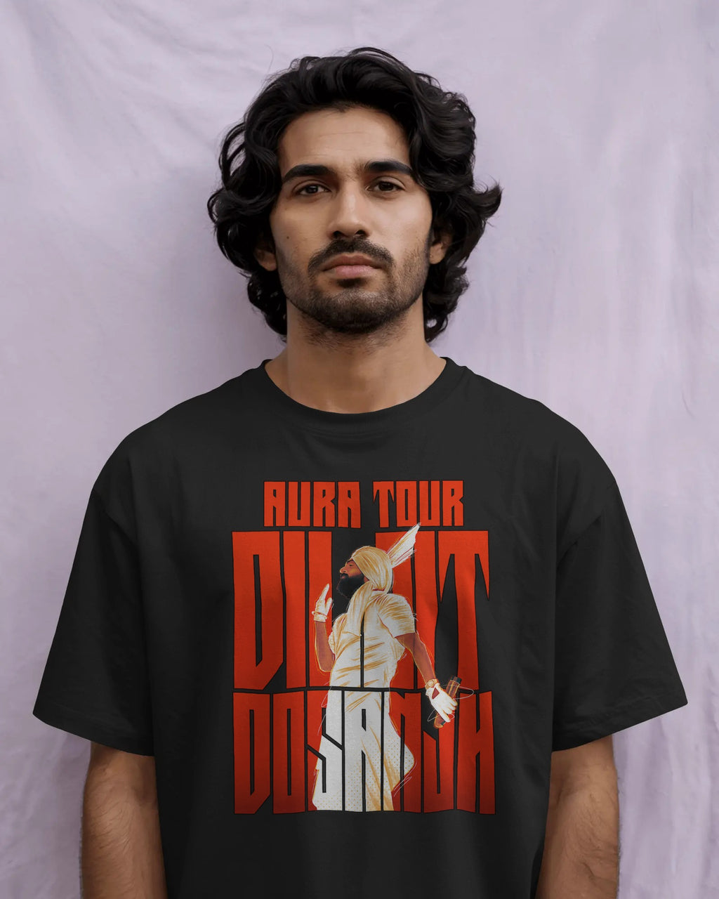 A man wearing a black t-shirt with a bold red and white graphic print that reads 'AURA TOUR Diljit Dosanjh' with an illustration of an Egyptian pharaoh, set against a light purple fabric backdrop.