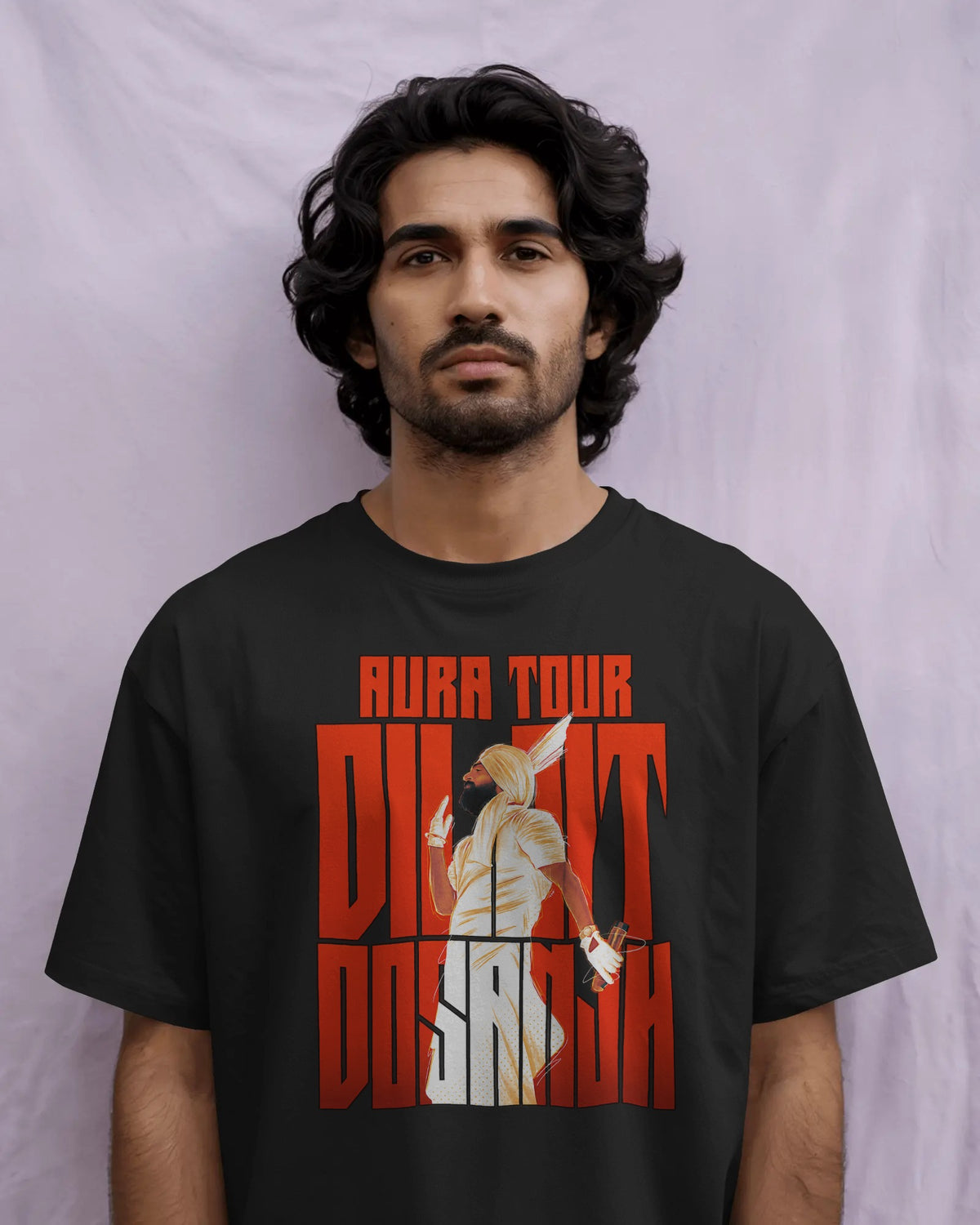 A man wearing a black t-shirt with a bold red and white graphic print that reads 'AURA TOUR Diljit Dosanjh' with an illustration of an Egyptian pharaoh, set against a light purple fabric backdrop.