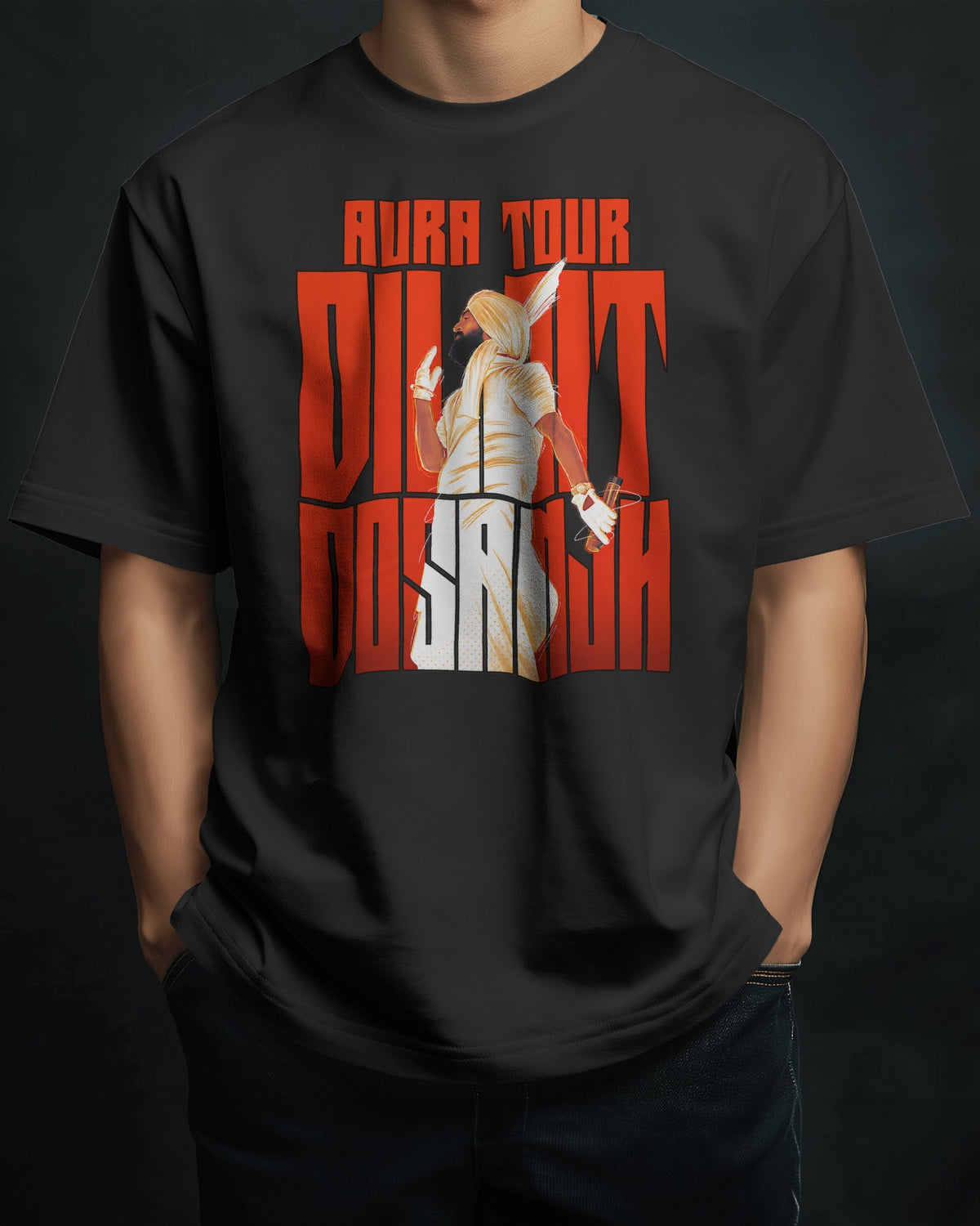 A person wearing a black graphic T-shirt with a large red, orange, and white 'Aura Tour Diljit Dosanjh Tshirt' print on the front, paired with dark jeans against a dark studio background.