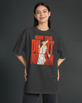 A woman modeling a dark charcoal gray oversized t-shirt with a large graphic print on the front. The graphic features the text 'AURA TOUR' in orange-red, 'Diljit Dosanjh in orange-red, with a stylized illustration of a woman in a white dress in the center.