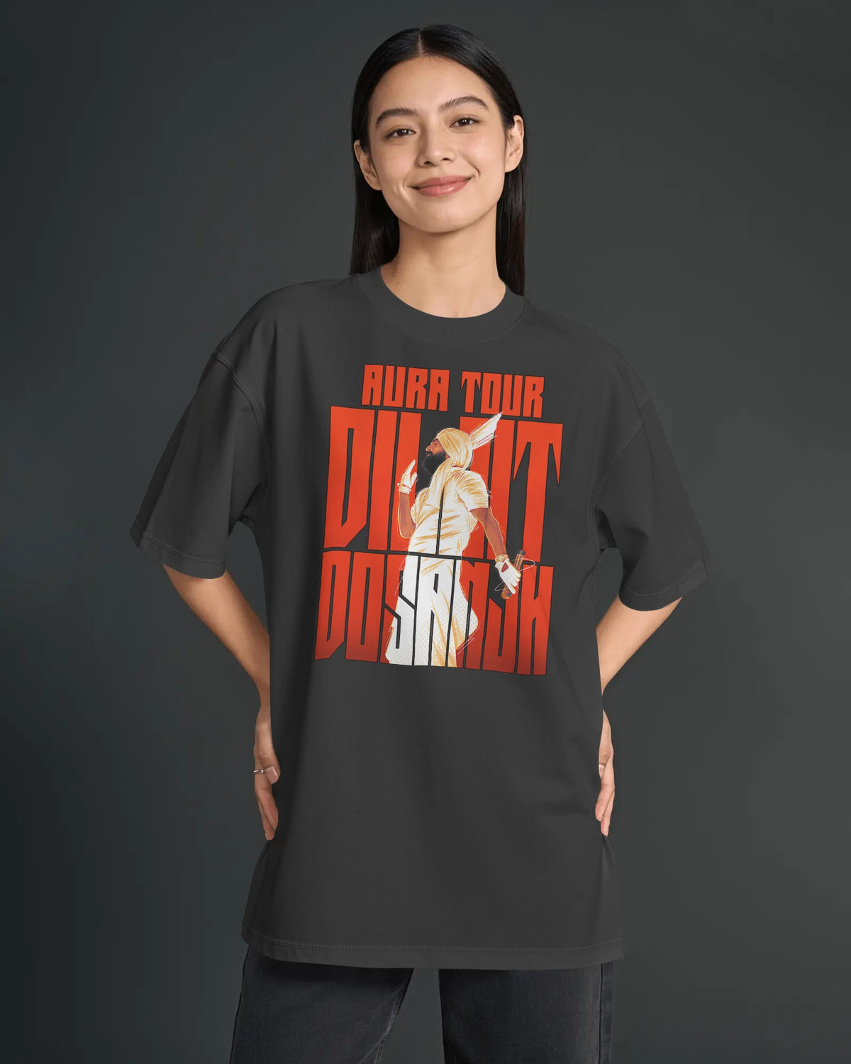 A woman modeling a dark charcoal gray oversized t-shirt with a large graphic print on the front. The graphic features the text 'AURA TOUR' in orange-red, 'Diljit Dosanjh in orange-red, with a stylized illustration of a woman in a white dress in the center.