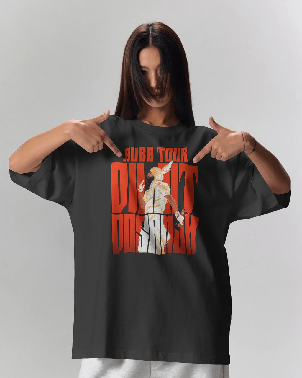 A woman wearing an oversized black graphic t-shirt with a red and white 'Diljit Dosanjh' print, pointing at the graphic with both hands against a light gray background.