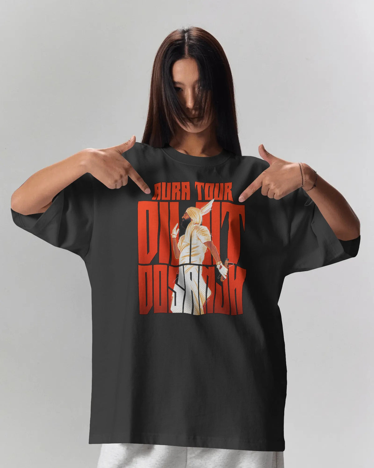 A woman wearing an oversized black graphic t-shirt with a red and white 'Diljit Dosanjh' print, pointing at the graphic with both hands against a light gray background.