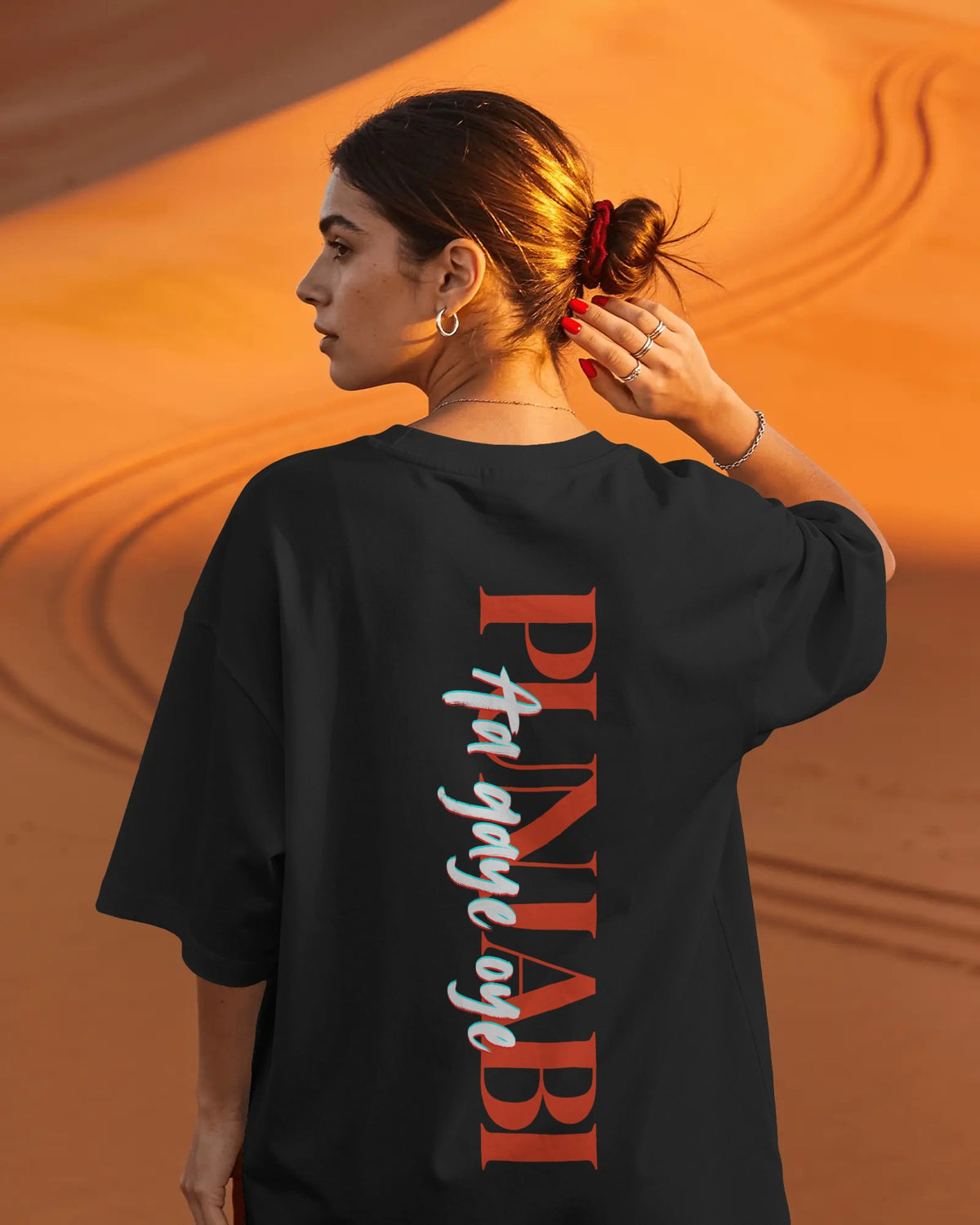 A woman photographed from behind wearing an oversized black T-shirt with large vertical red, white, and blue text on the back, standing against a warm orange desert dune background. Diljit Dosanjh Aura Tour India Tshirt