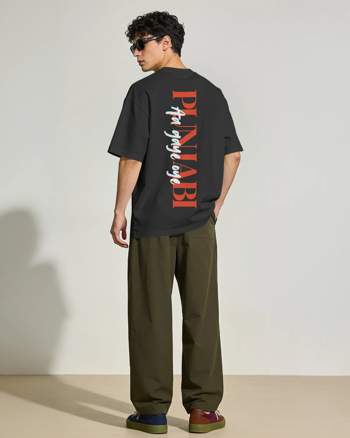 A man wearing a black short-sleeve t-shirt with a large red and white vertical graphic on the back, paired with olive green pants and sunglasses, standing against a plain light beige studio background. Diljit Dosanjh Punjabi aa gaye oye tshirt