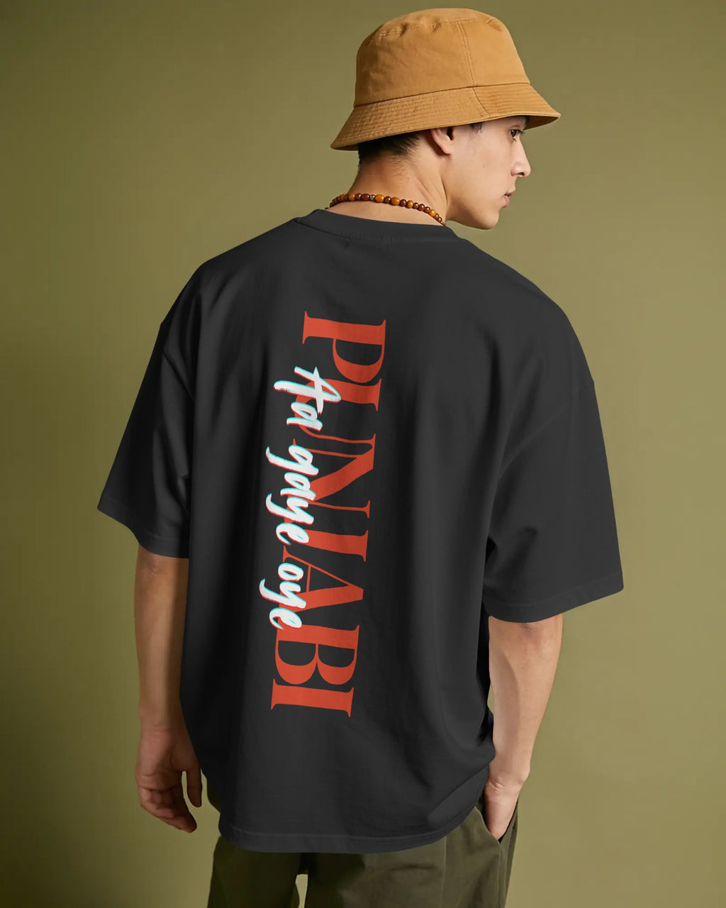 A man wearing a black oversized t-shirt with a large red and white graphic print on the back, paired with a tan bucket hat and olive pants, shown from behind against a solid olive green background.