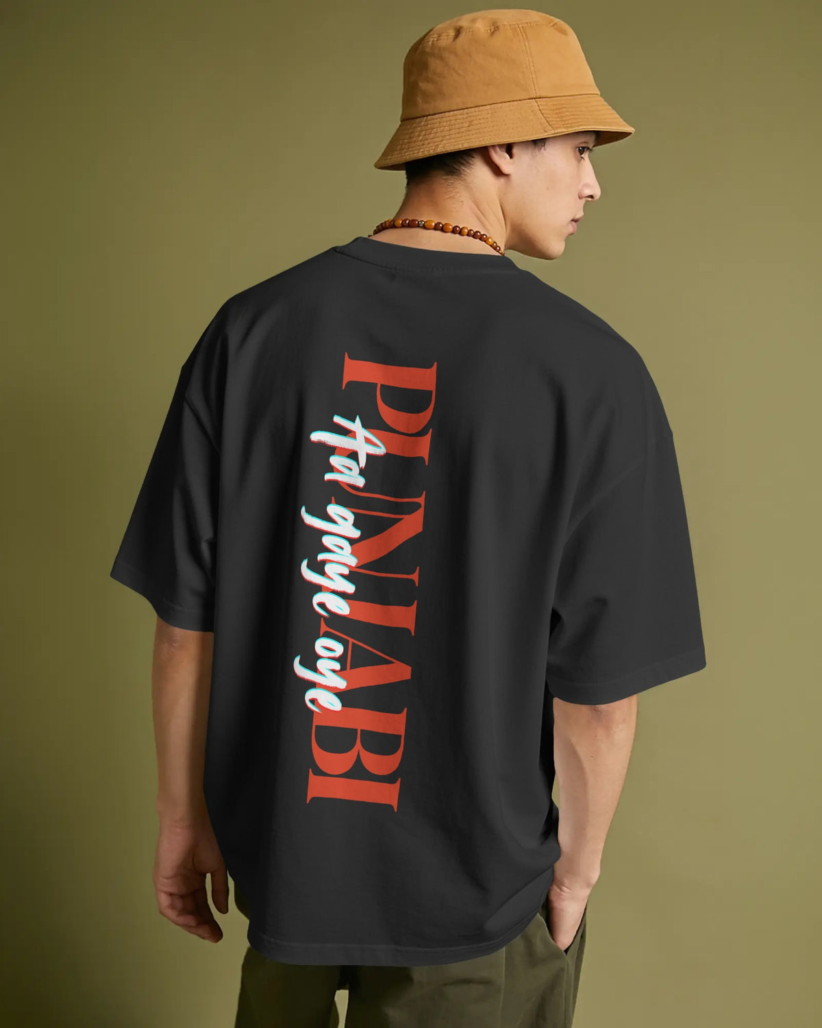 A man wearing a black oversized t-shirt with a large red and white graphic print on the back, paired with a tan bucket hat and olive pants, shown from behind against a solid olive green background.