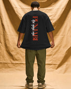 A man stands with his back to the camera wearing an oversized black t-shirt with a vertical red and white graphic print on the back, paired with olive green pants and beige sneakers, set against a draped tan fabric backdrop. Punjabi aa gaye oye