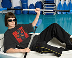 A model wearing a black graphic t-shirt with a red and yellow Nitorious Atelier Diljit Dosanjh Aura Tour Collection. design, black pants with yellow piping, and sunglasses, reclining in a white kayak while holding a blue paddle in a pool with kayaks in the background.