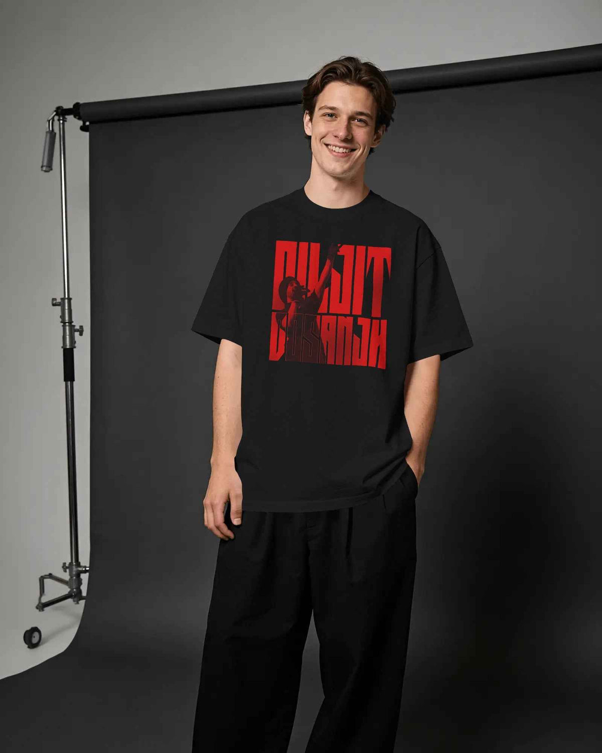 A man wearing a black oversized t-shirt with a bold red graphic print that reads 'Diljit Dosanjh Aura Tour' in a stylized font, paired with black trousers, standing in front of a dark gray studio backdrop. Nitorious Atelier Diljit Dosanjh Aura Tour Collection.