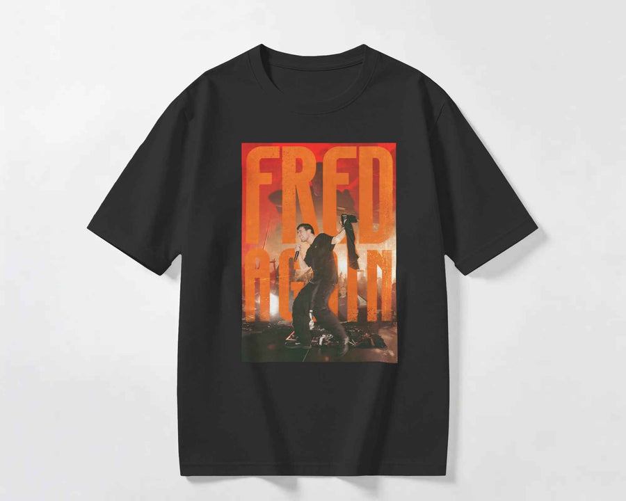 A black short-sleeve crewneck t-shirt featuring a large graphic print on the front with the text 'FRED AGAIN' in orange and yellow over a photo of a man playing a guitar. Fred Again India Concert collection