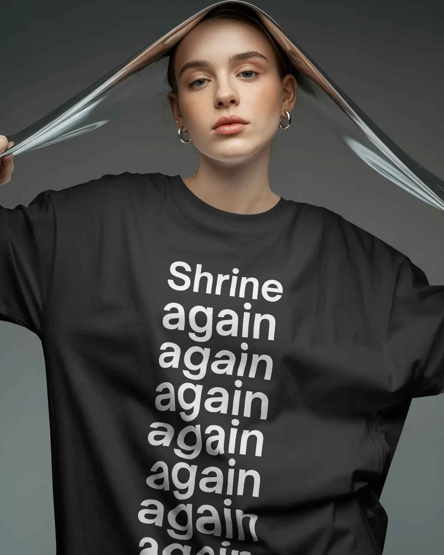 A model wearing a black oversized t-shirt with the word 'Shrine' repeated in white text, holding a clear plastic sheet overhead against a dark gray studio background. Fred Again India Concert collection