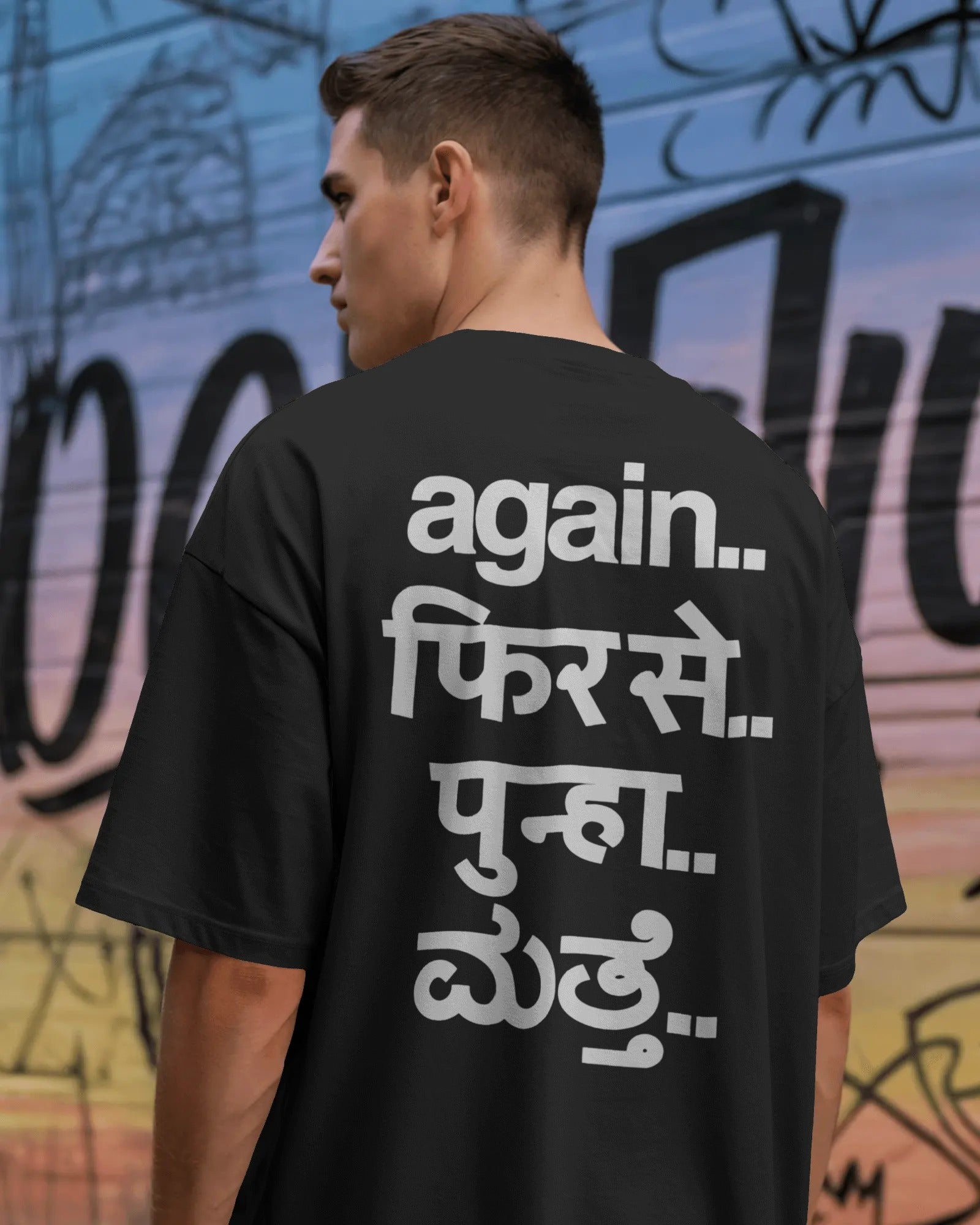 A man viewed from behind wearing a black t-shirt with white Hindi text printed on the back, set against a colorful graffiti wall. Fred Again India concert collection Tshirt 
