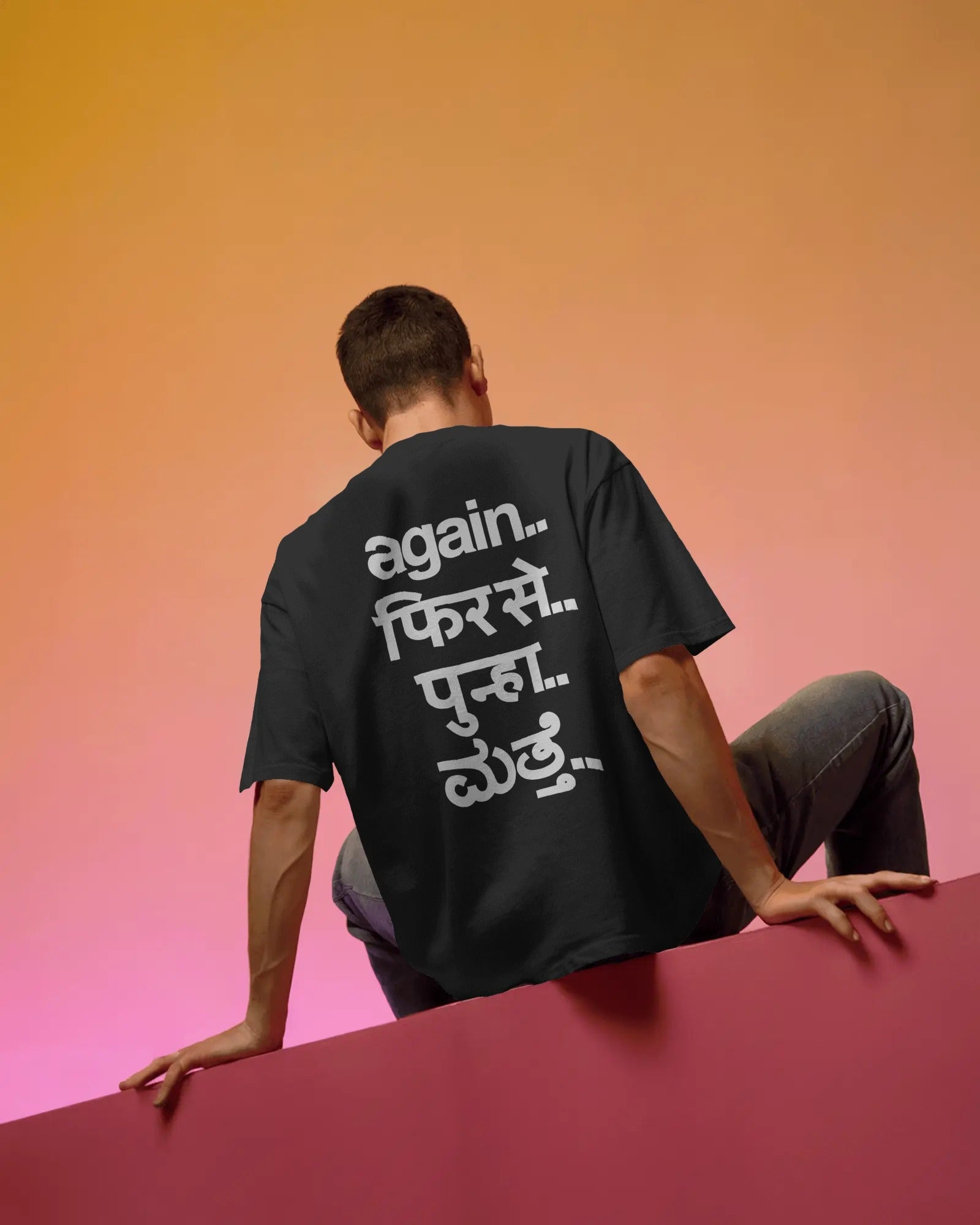 A man wearing a black t-shirt with white Hindi text on the back, sitting on a pink platform against a warm orange-to-pink gradient background. Fred Again.. Tshirt India