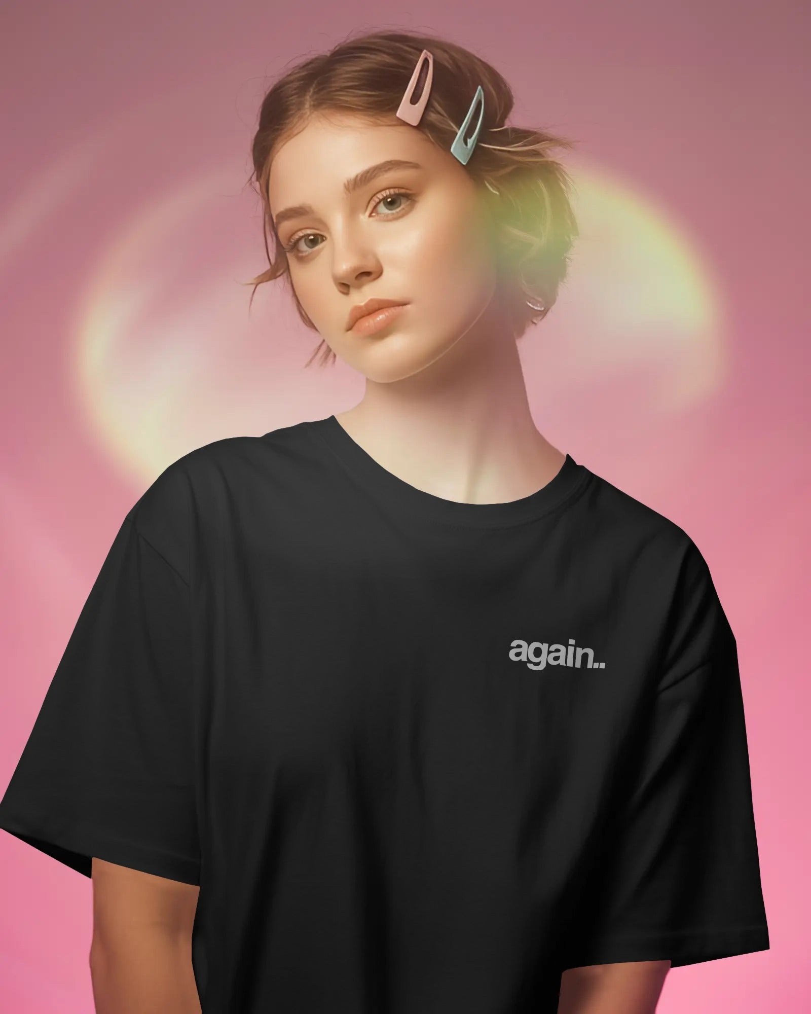 A woman wearing an oversized black t-shirt with the word 'again.' printed in white on the left chest, posed against a pink background with a soft green glow.