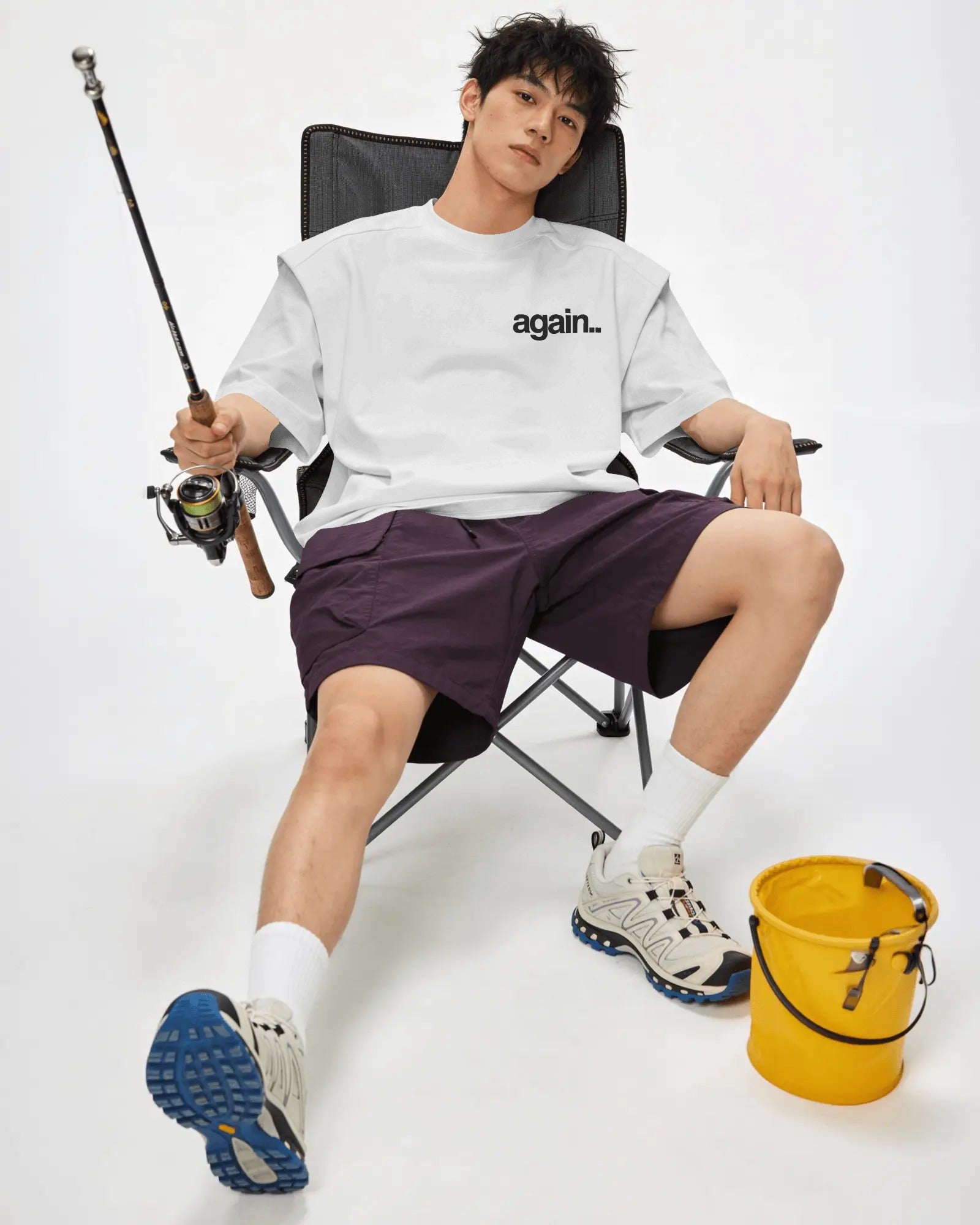 A man sits in a black folding camp chair wearing a white t-shirt with 'again. Fred Again.. Tshirt India