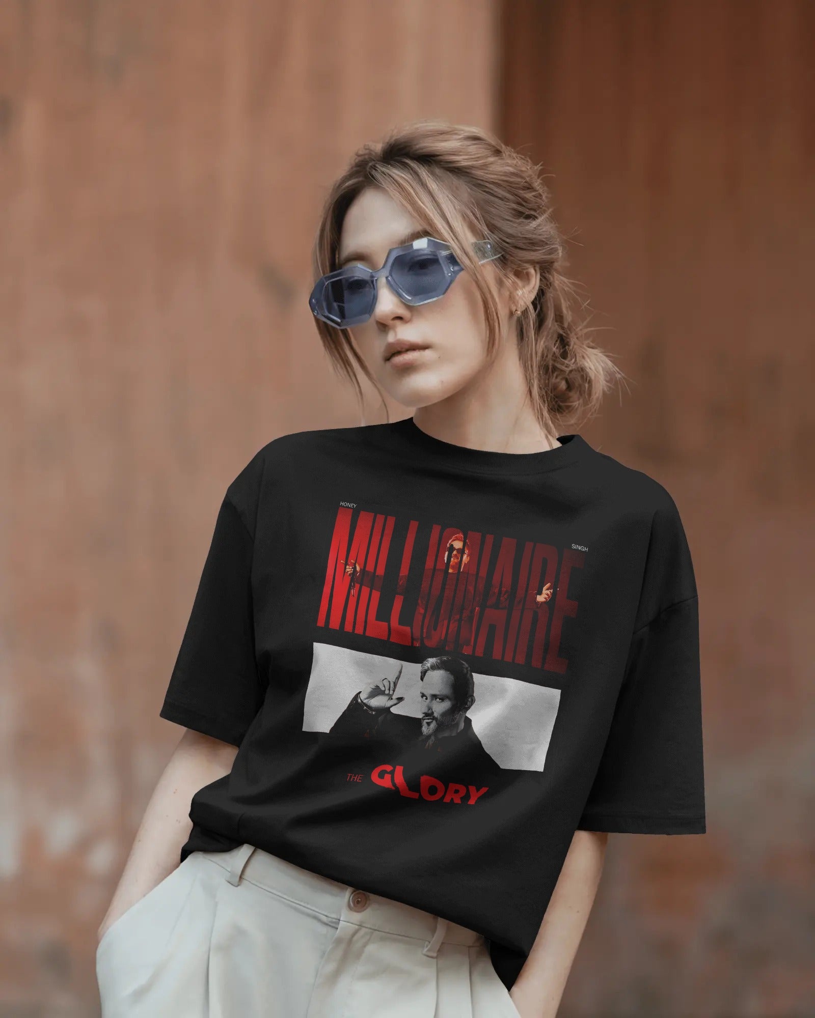 Person wearing a black t-shirt with 'Millionaire Glory' graphic design against a textured brown wall.