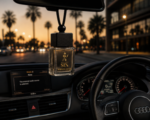 A clear glass car air freshener bottle labeled 'Silent Sin Morning Affair' hangs from a black string under the rearview mirror inside a car at sunset, with the dashboard, steering wheel, and blurred palm trees outside visible.