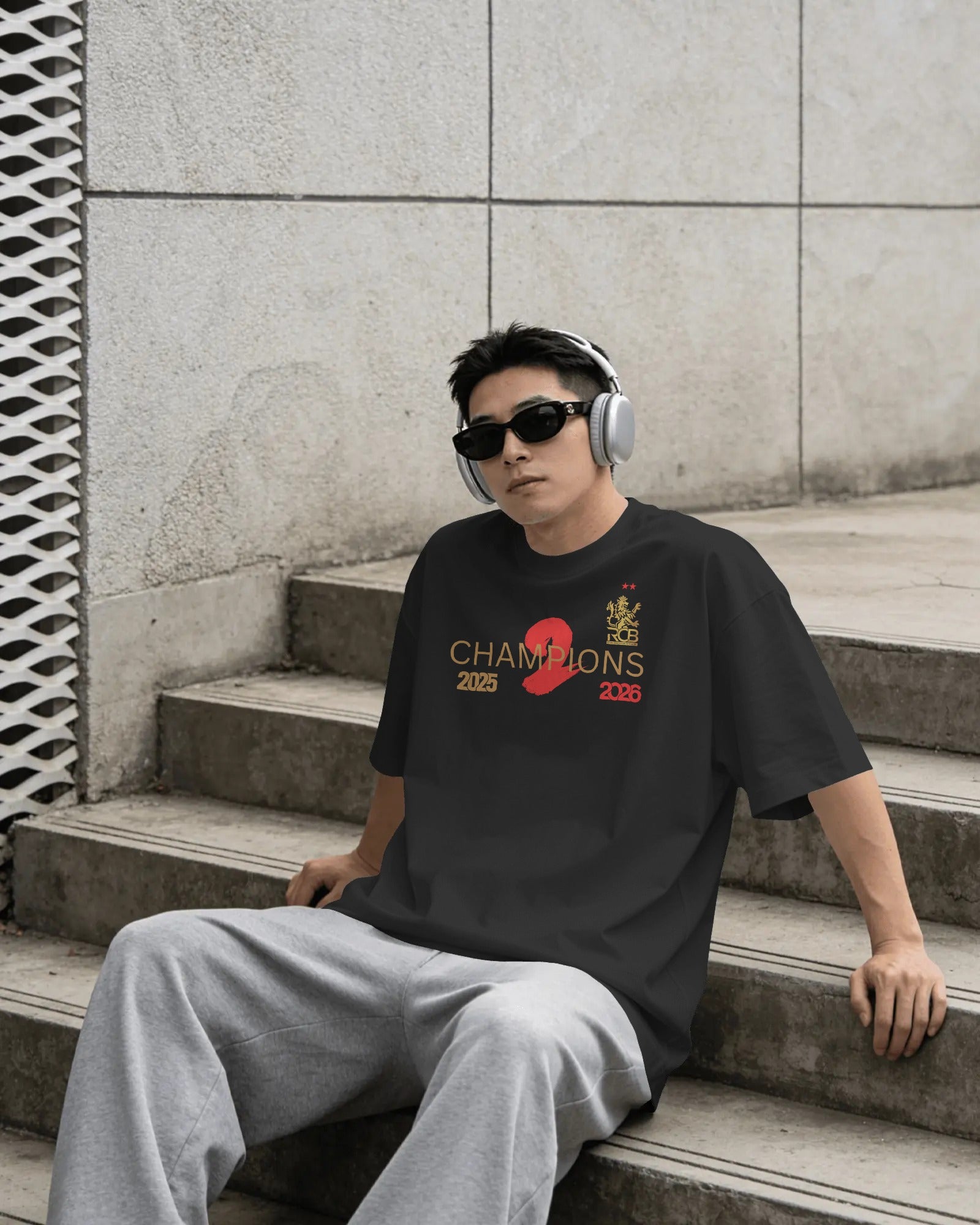 Person wearing a black t-shirt with 'Champions 2026' text, sitting on steps outdoors. RCB Champions Tshirt from Nitorious Atelier 