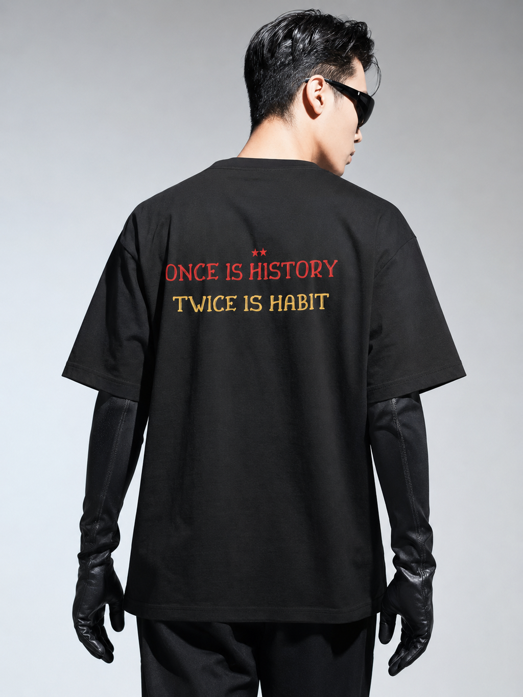 Person wearing a black t-shirt with text on the back, standing against a gray background