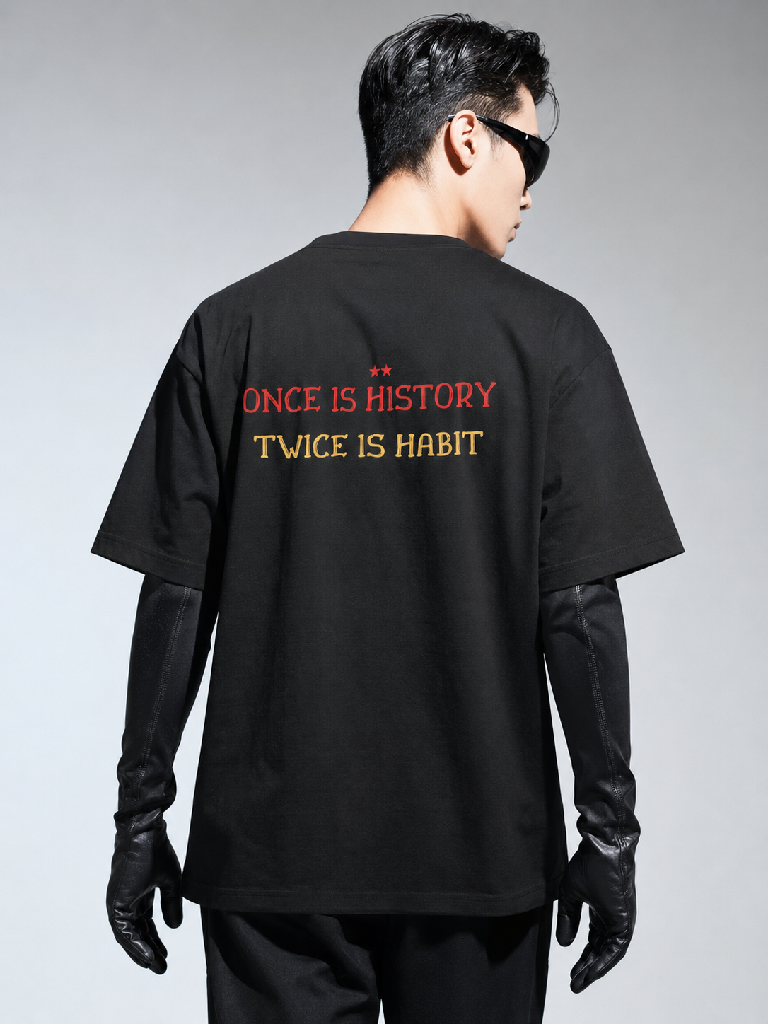 Person wearing a black t-shirt with text on the back, standing against a gray background