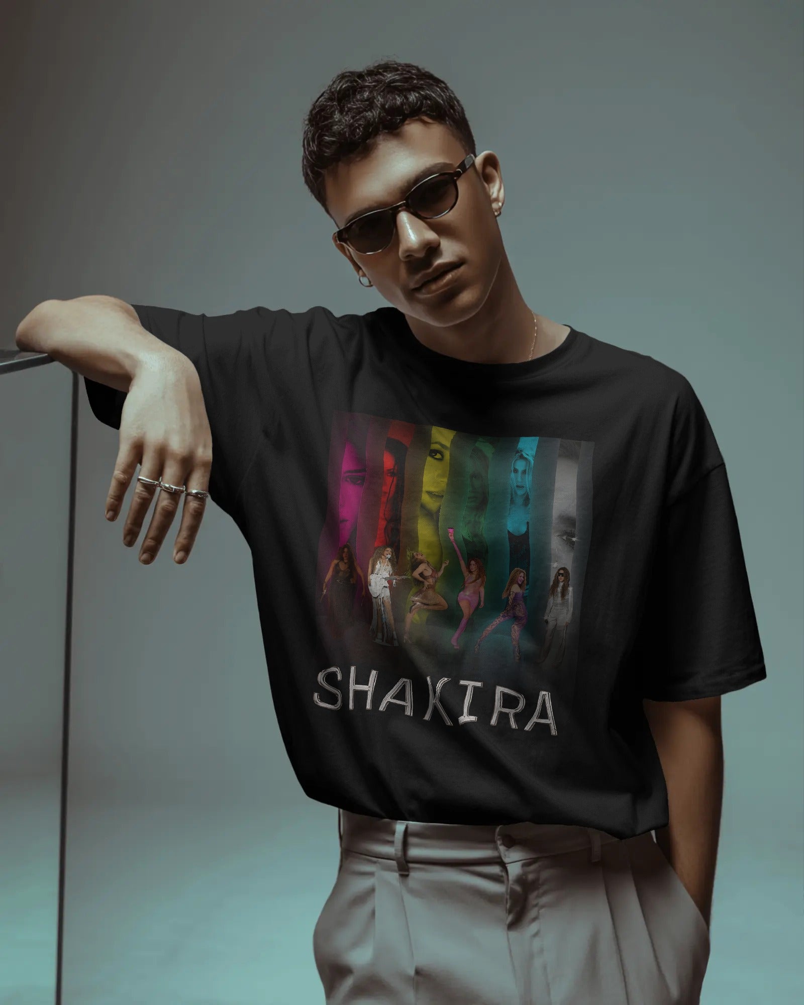 Person wearing a black t-shirt with a colorful graphic and 'Shakira' text, leaning against a gray background.