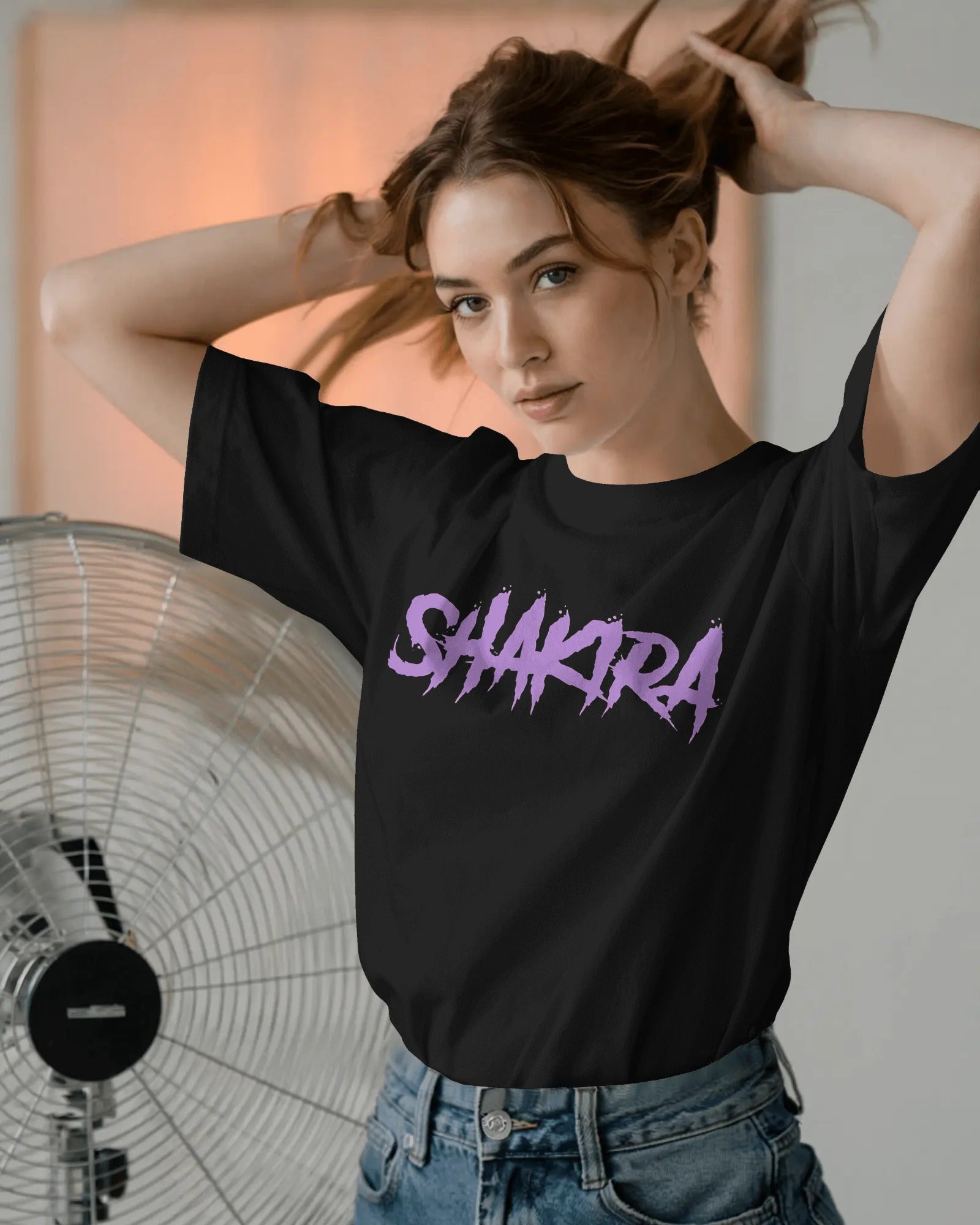 Woman wearing a black t-shirt with 'SHAKIRA' printed in purple, standing in front of a fan.