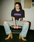 Person wearing a black t-shirt with 'SHAKIRA' text, ripped jeans, and brown boots sitting on a chair.