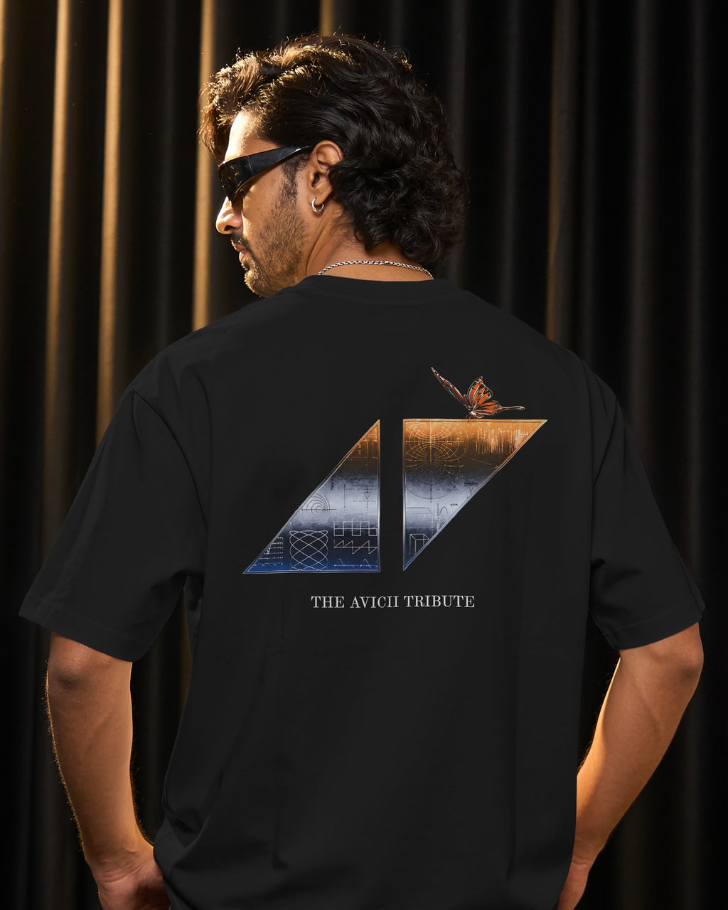A man wearing a black t-shirt from the back, featuring a large graphic print with a butterfly and the text 'THE AVICII TRIBUTE' on a dark curtain background.
