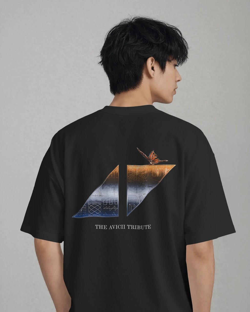 A man wearing a black short-sleeve t-shirt, viewed from the back. The shirt features a large graphic print on the back depicting a split landscape with a butterfly and the text 'THE AVICII TRIBUTE'.