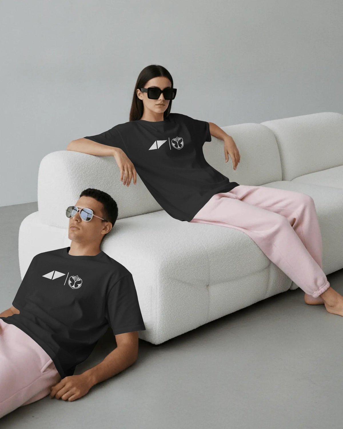 Two models wearing matching black graphic t-shirts and light pink sweatpants, posed on a modern white textured sofa in a minimalist studio setting. The Avicii Tribute Tomorrowland Tshirt.