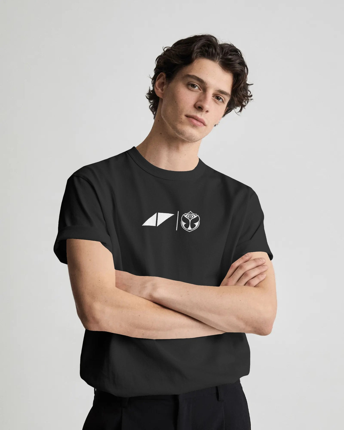 A man wearing a black short-sleeve crewneck t-shirt with a white printed logo on the chest, posed with arms crossed against a light gray studio background.