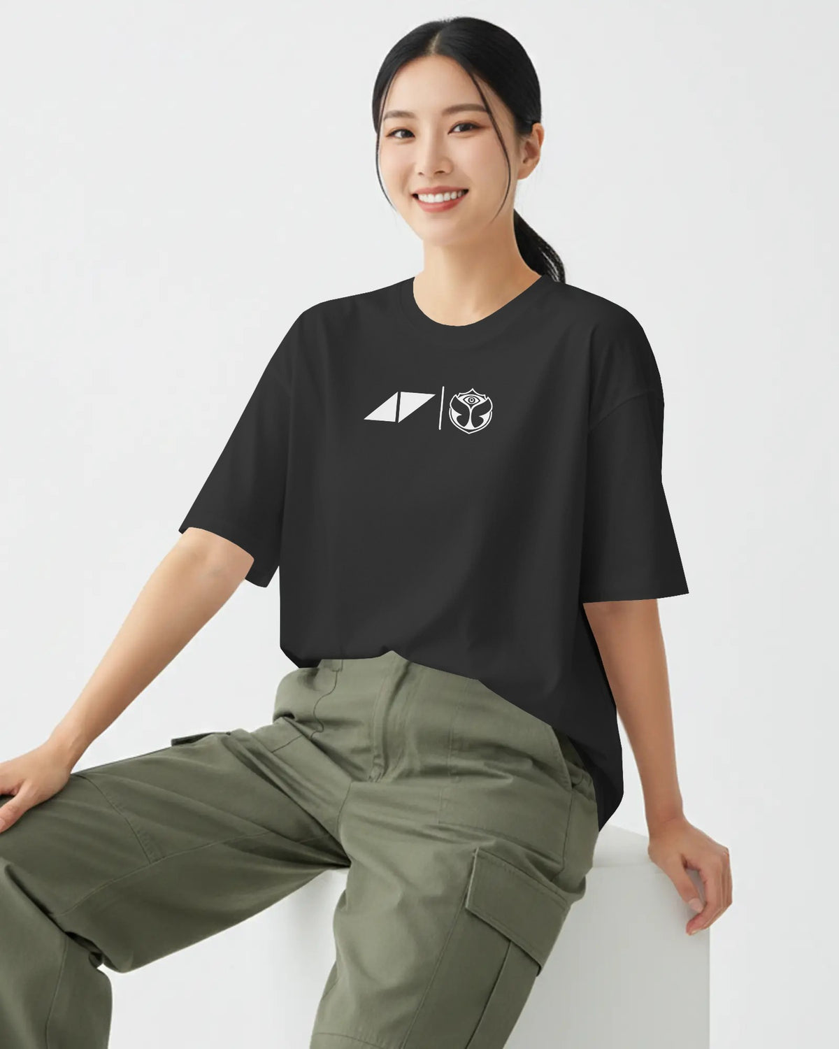 A woman wearing a black short-sleeve t-shirt with a white geometric chest graphic and olive green cargo pants, sitting against a clean white background.