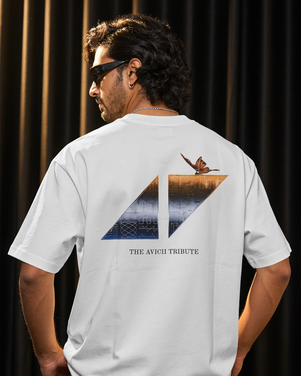 A man wearing a white t-shirt from the back, featuring a large graphic print with a butterfly, a split mountain design, and the text 'THE AVICII TRIBUTE' against a dark curtain background.
